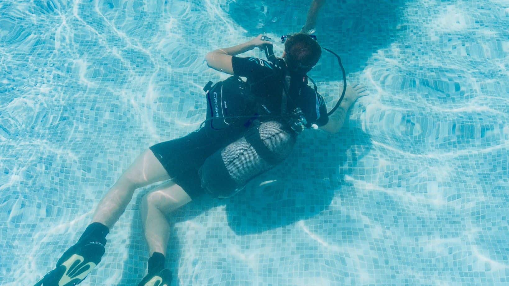 Scuba diver with full gear floating underwater in a bright blue swimming pool.