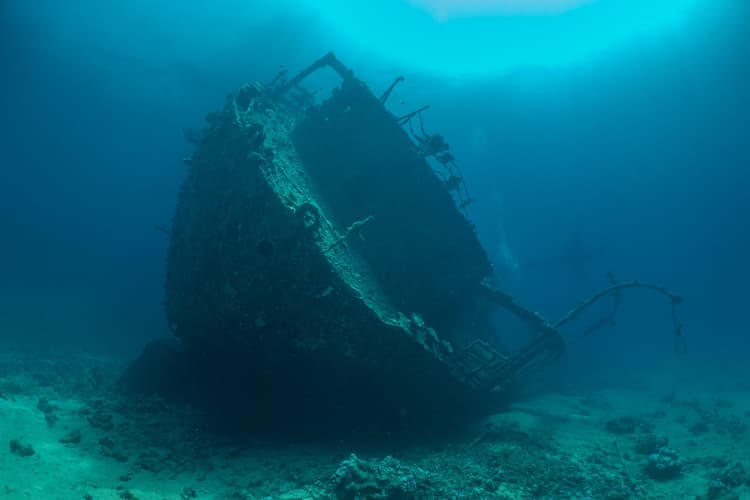 A large, decaying shipwreck resting on the sandy seabed, with sunlight illuminating the water.