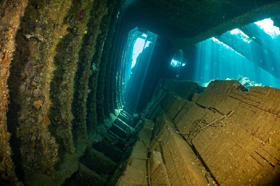 A diver exploring the cavernous interior of a large shipwreck, with light entering from several openings.