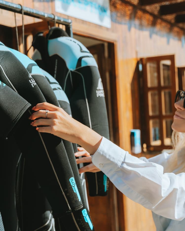 Person's hand reaching to touch a black and blue wetsuit hanging on a rack.