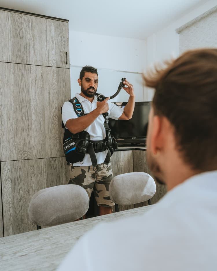 A dive instructor demonstrating scuba gear components to another man in an indoor setting.