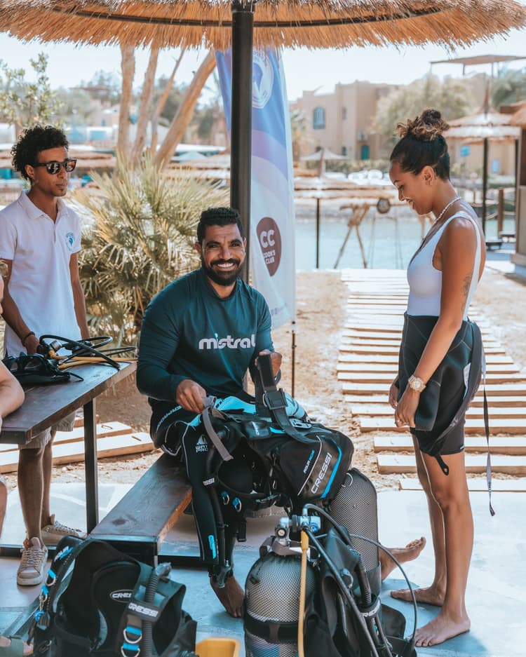 Smiling dive instructor assisting two students with their scuba gear outdoors.