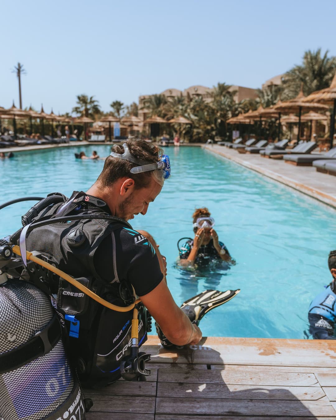 A diver preparing equipment by the edge of a pool, with another diver already in the water.