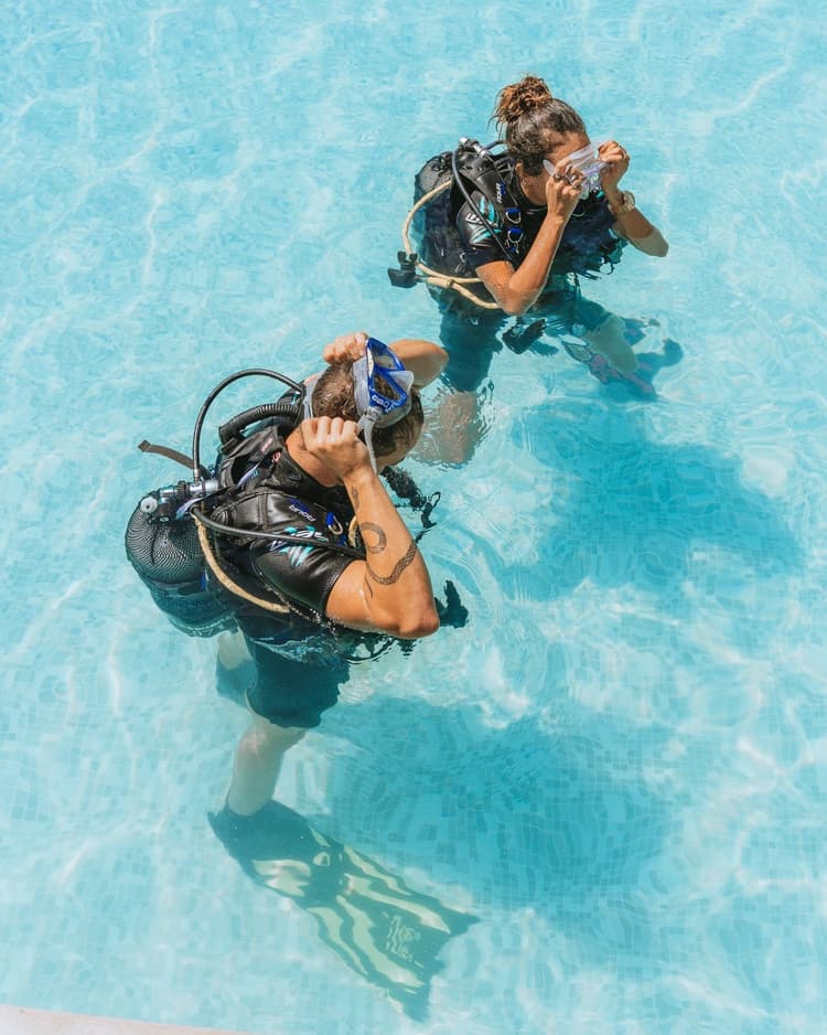 Overhead view of two scuba divers adjusting masks in a clear blue pool.