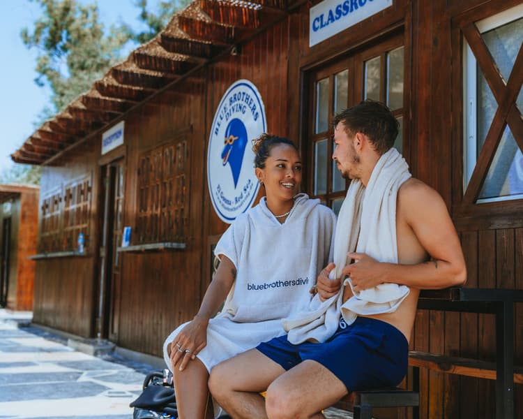 A couple in swimsuits sitting outside a wooden dive center building, smiling at each other.