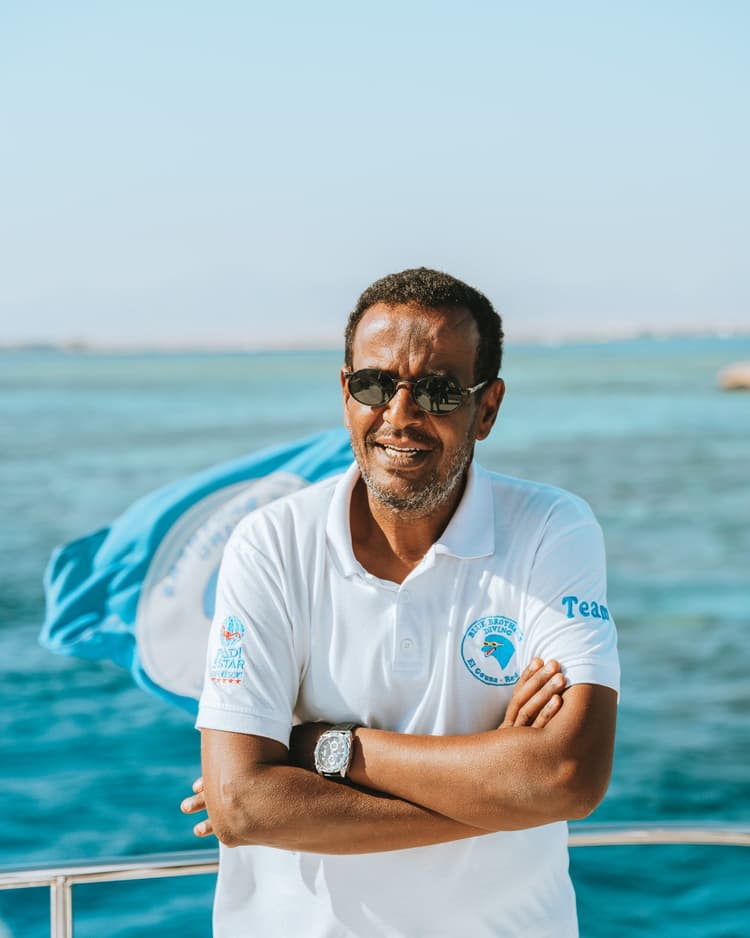 A man in sunglasses and a white polo shirt with a blue logo, arms crossed, standing on a boat.