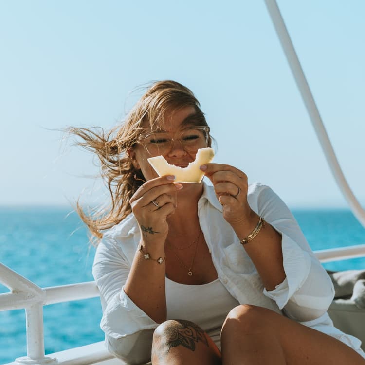 Woman smiling, eating a snack on a boat with blue ocean and sky.