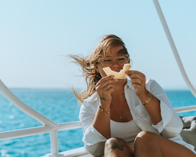 A woman laughing heartily and holding a slice of fruit on a boat deck.