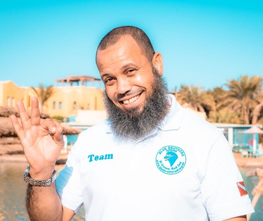 Bearded man in white polo shirt smiles, making an OK gesture outdoors.