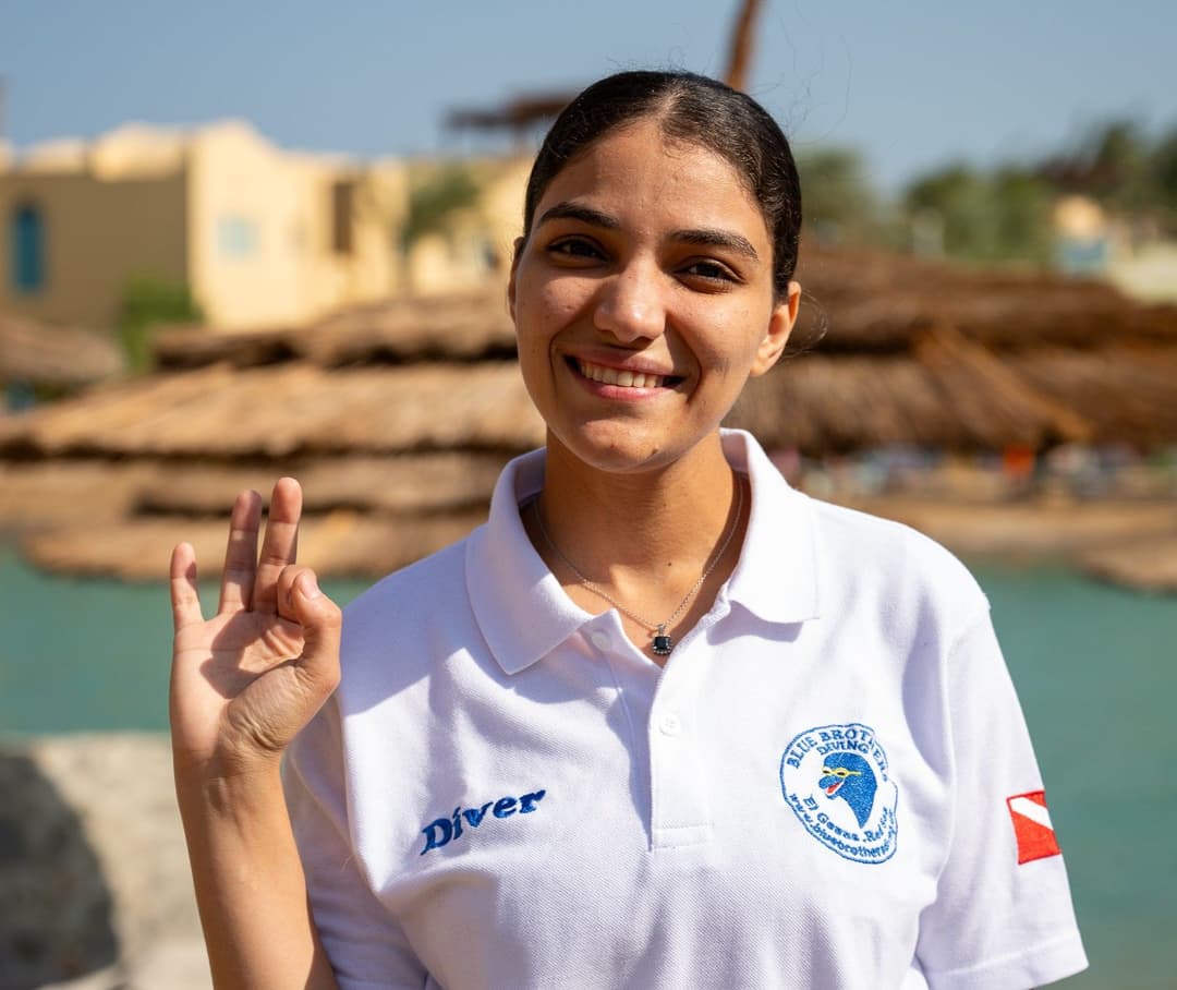 Young woman in white dive polo shirt making an 'ok' sign outdoors.
