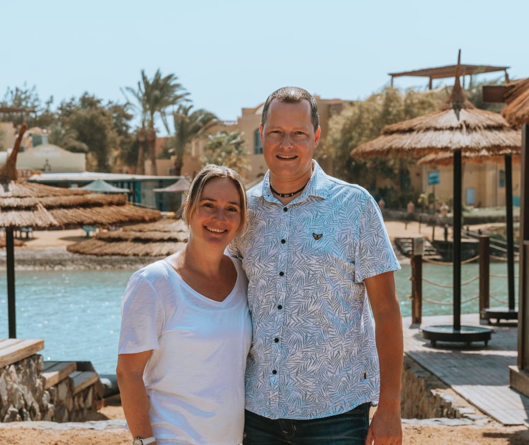 Happy couple stands by tranquil blue water with resort buildings and straw umbrellas.