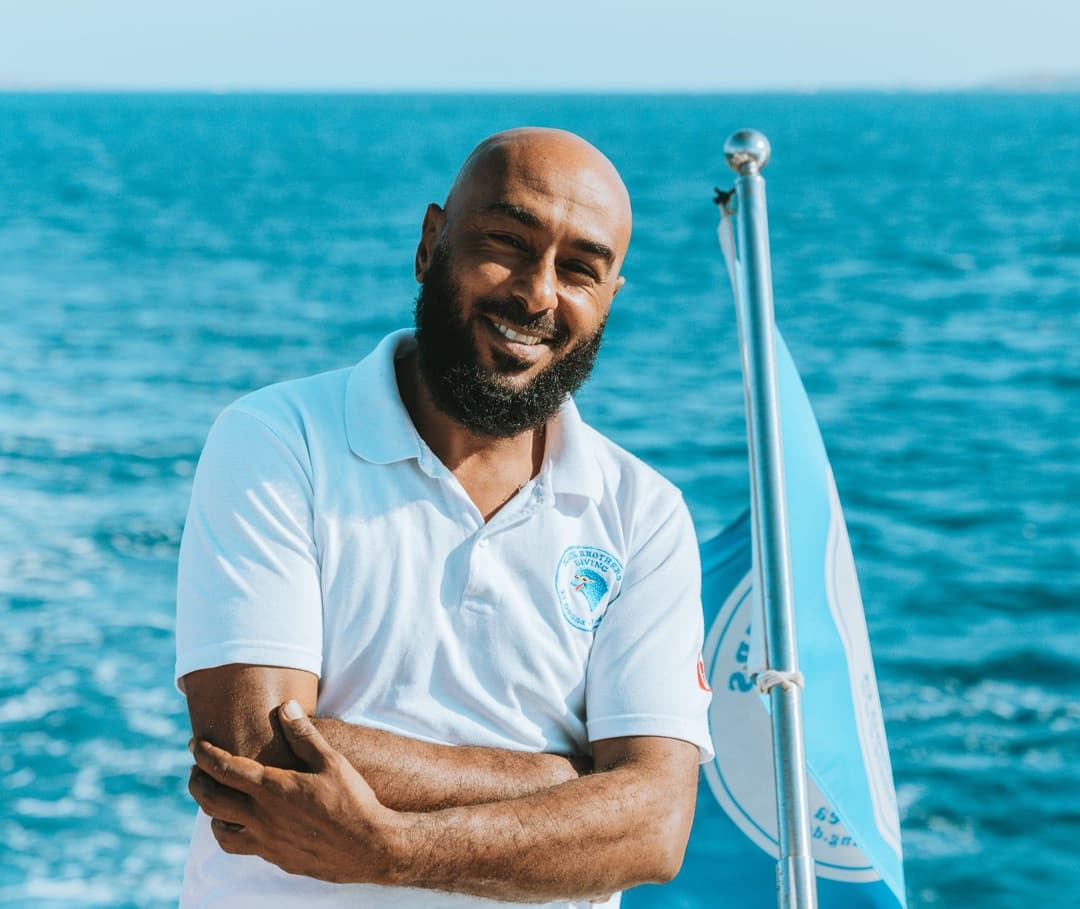 Bearded man in white polo shirt smiles with crossed arms on a boat.