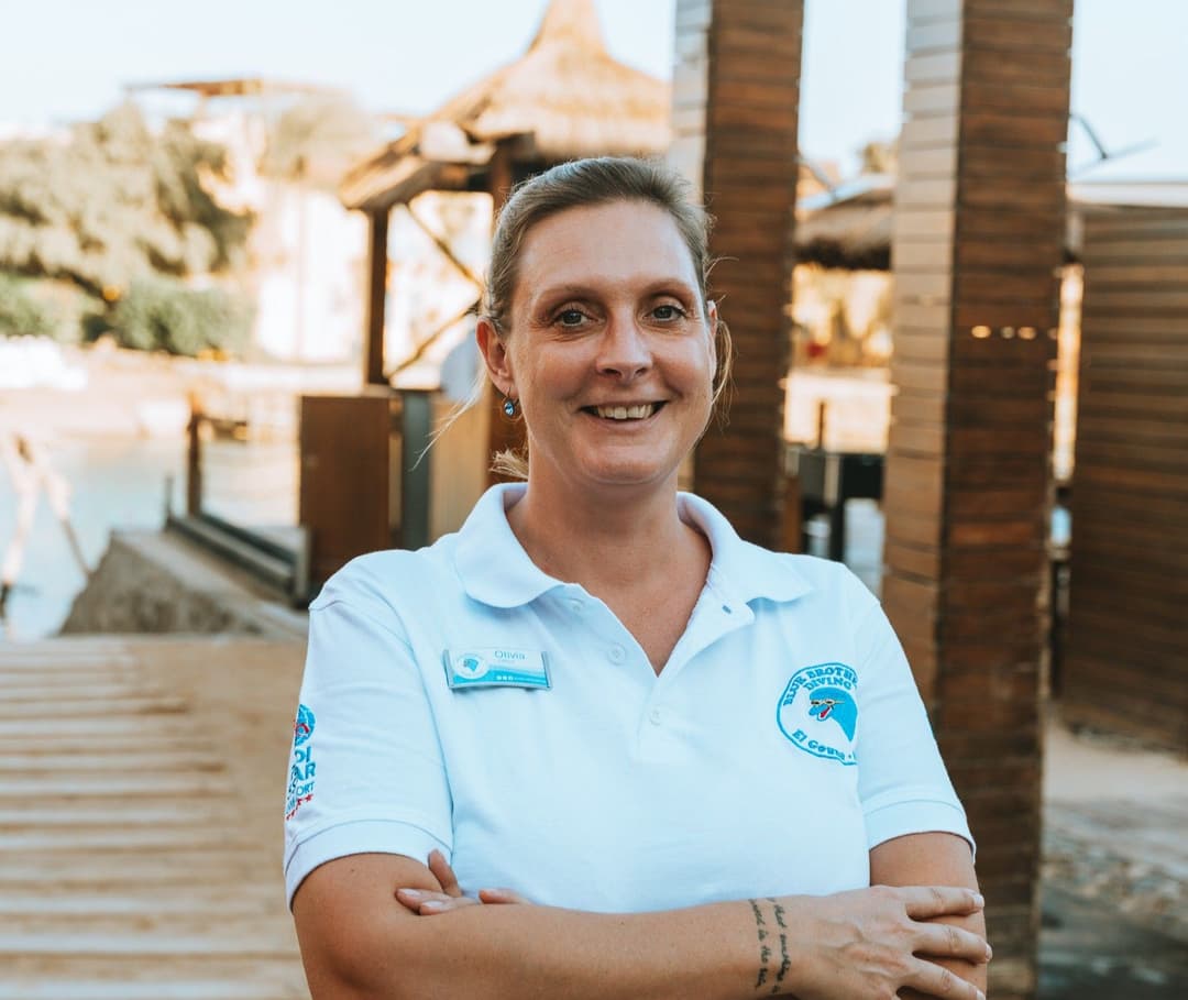 Smiling woman in white polo shirt with logos, arms crossed, outdoor setting.