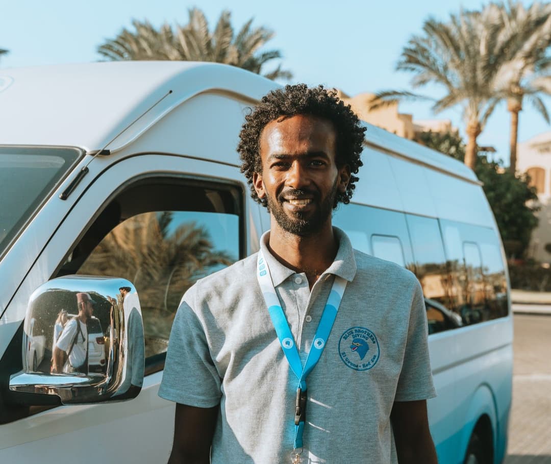 Smiling man in a grey polo shirt standing by a white tour van.