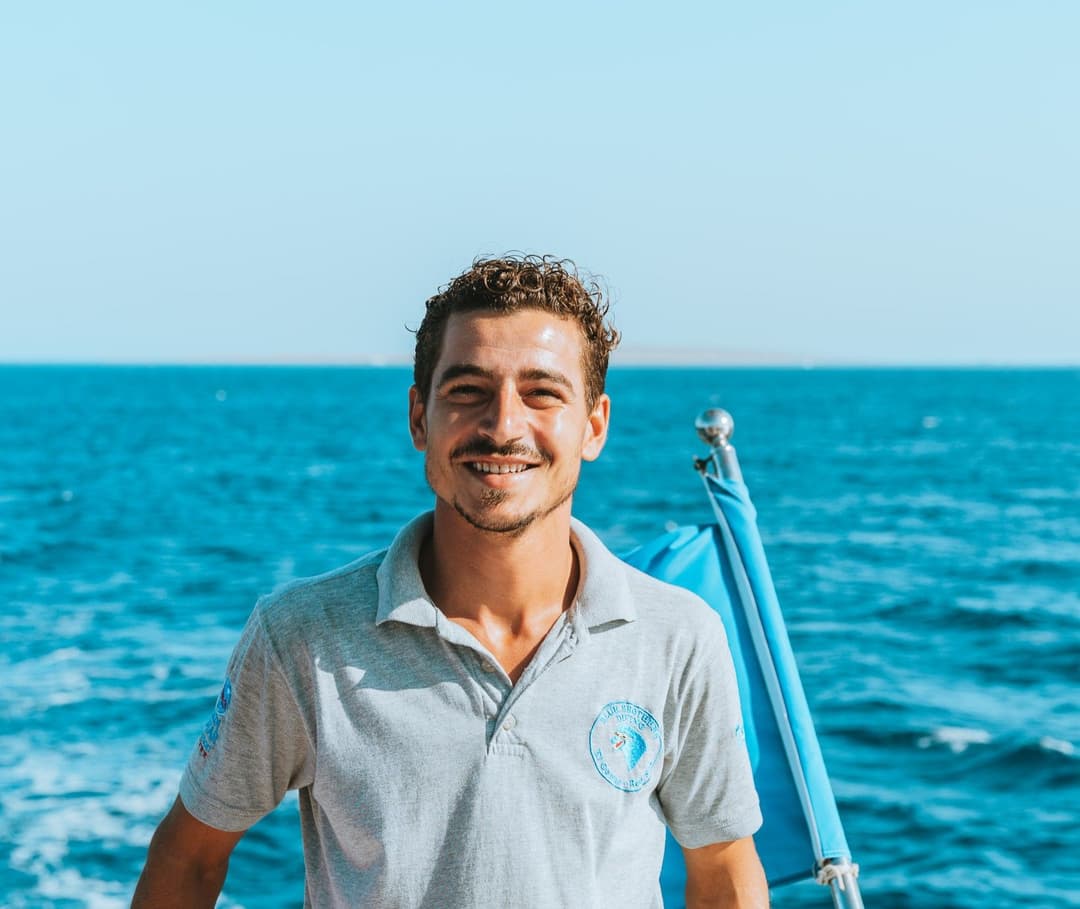 Smiling man in grey polo shirt on a boat with blue ocean and sky background.