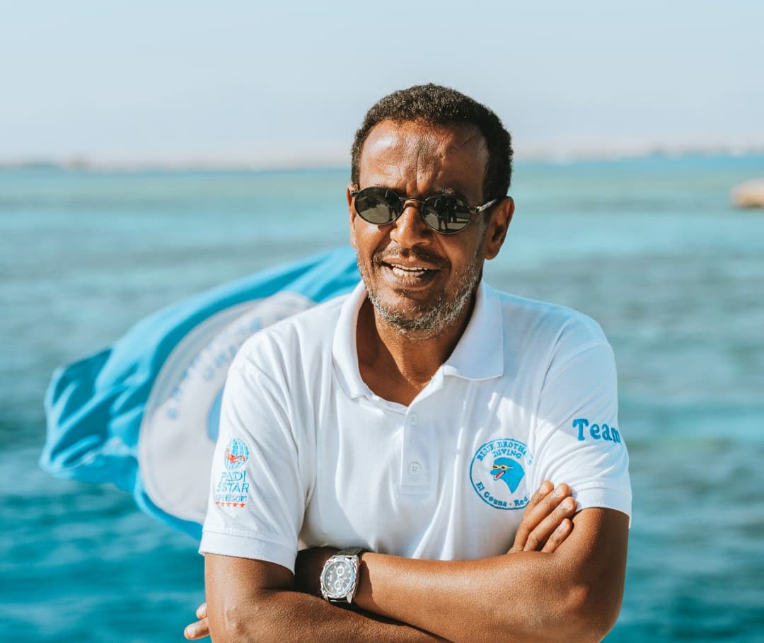 Man in sunglasses, white polo shirt, arms crossed, standing by the blue ocean.