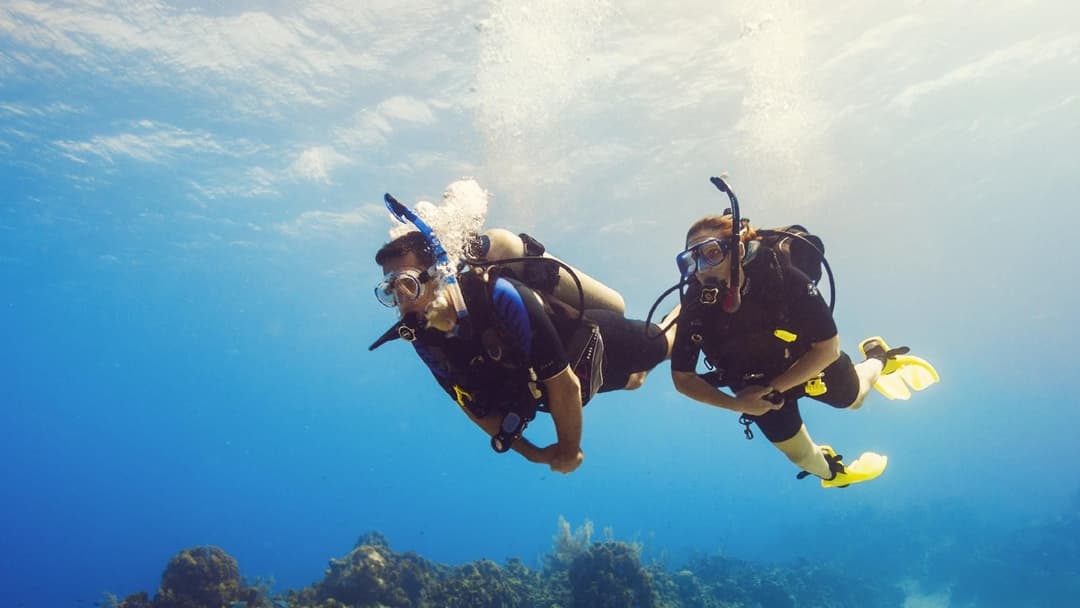 Two scuba divers swim in clear blue ocean water with sunlight filtering from above.